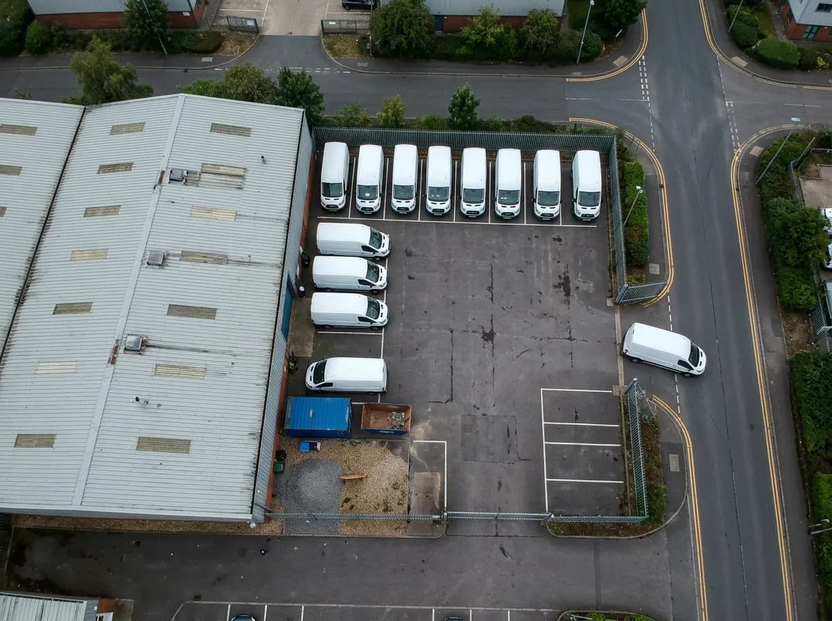 Overhead view of a depot yard with a row of white vans, and one van driving out onto the road